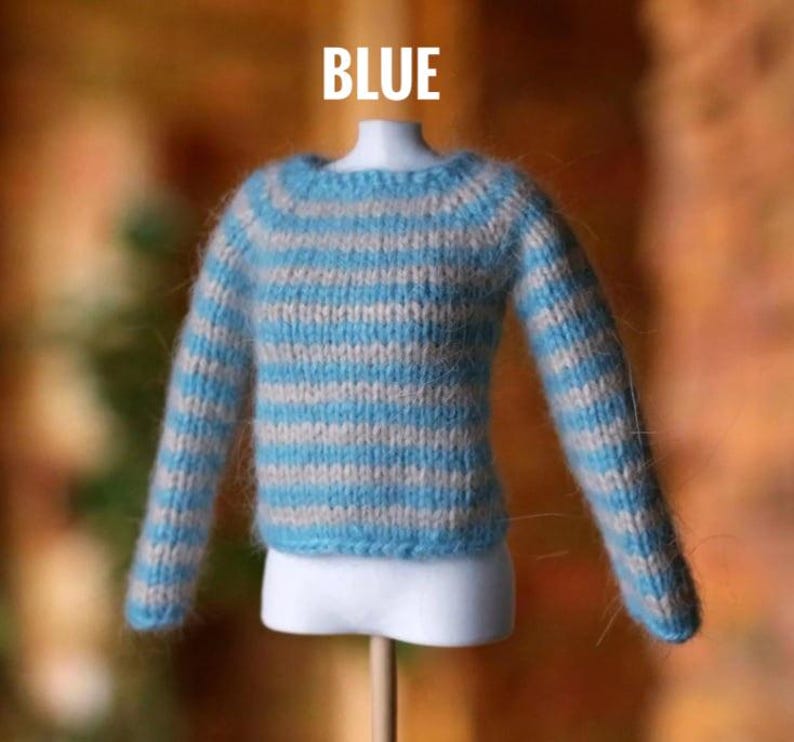 May include: A blue and light gray striped sweater for a doll. The sweater is made of a soft, fuzzy material and has long sleeves. The word "BLUE" is written in white block letters above the sweater.