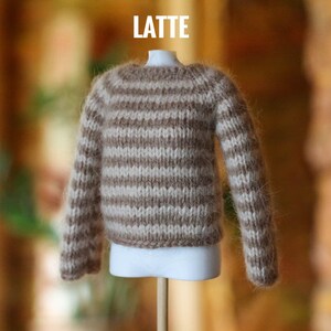 May include: A miniature, hand-knitted sweater in a latte color scheme. The sweater features horizontal stripes of light beige and brown. The word "LATTE" is displayed in white block letters at the top. The sweater is displayed on a white mannequin.