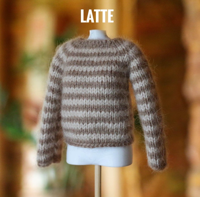 May include: A miniature, hand-knitted sweater in a latte color scheme. The sweater features horizontal stripes of light beige and brown. The word "LATTE" is displayed in white block letters at the top. The sweater is displayed on a white mannequin.