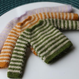 May include: Close-up of four knitted items in various colors: lavender, orange, and green. Each item has horizontal stripes. The items are arranged on a white surface, with a teal background.