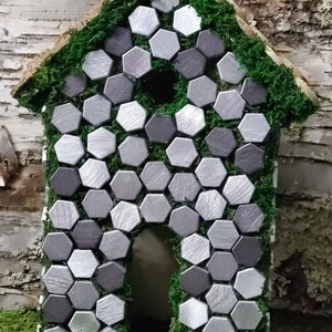 May include: A small house-shaped birdhouse made of wood and covered in moss. The front of the house is decorated with silver and gray hexagonal tiles.