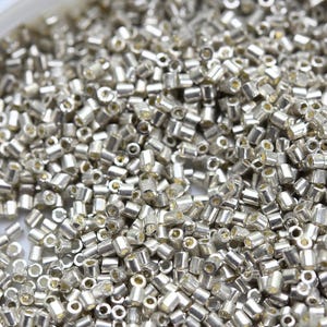 May include: Close-up of a pile of small, silver-colored seed beads. The beads are cylindrical with a hole running through the center, and they have a metallic sheen. The beads are scattered, creating a textured surface.