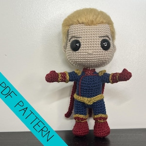 May include: A crocheted doll featuring a large head with blonde hair and black eyes, wearing a blue suit with gold accents, red gloves, red boots, and a red cape. A diagonal blue banner across the bottom left corner displays the text PDF PATTERN.