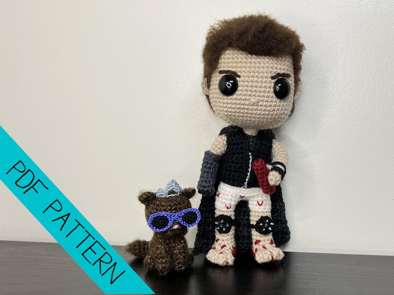 May include: Crocheted doll of a character with a black vest, cape, and white pants with red accents. A small brown crocheted dog with blue glasses and a crown is also present. The words "PDF PATTERN" are in a diagonal blue banner.