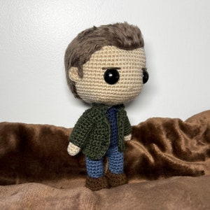 May include: A crocheted doll with brown hair, a green jacket, blue jeans, and brown shoes. The doll is sitting on a brown fabric surface.