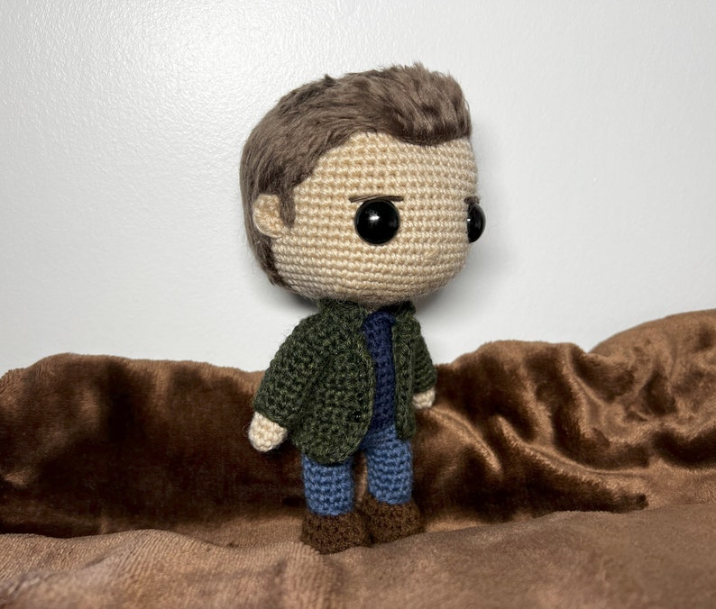 May include: A crocheted doll with brown hair, a green jacket, blue jeans, and brown shoes. The doll is sitting on a brown fabric surface.