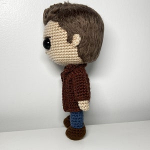 May include: Crocheted doll with brown hair, a brown jacket, blue jeans, and brown shoes. The doll is standing on a white surface.