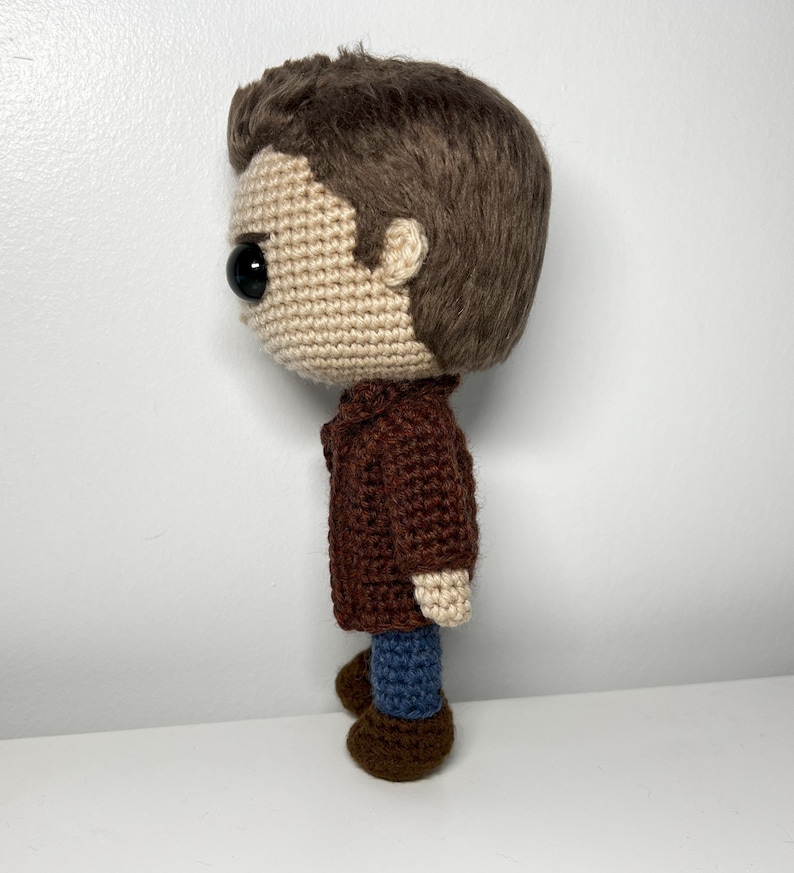 May include: Crocheted doll with brown hair, a brown jacket, blue jeans, and brown shoes. The doll is standing on a white surface.
