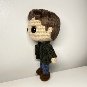 May include: A crocheted doll with brown hair, a green jacket, blue jeans, and brown shoes. The doll has large black eyes and a neutral expression.