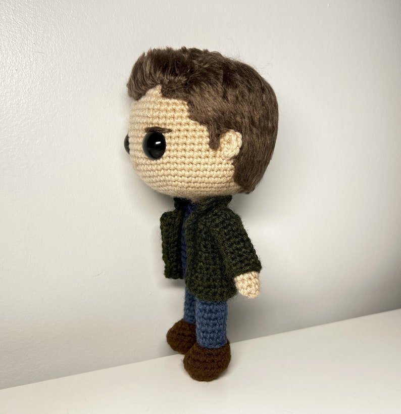 May include: A crocheted doll with brown hair, a green jacket, blue jeans, and brown shoes. The doll has large black eyes and a neutral expression.