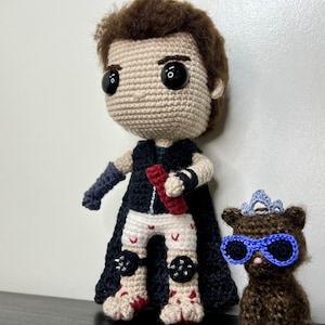 May include: Crocheted doll of a character with black cape, black and white pants, and a red object. Next to it is a crocheted bear wearing blue glasses and a crown. The dolls are handmade and have large black eyes.