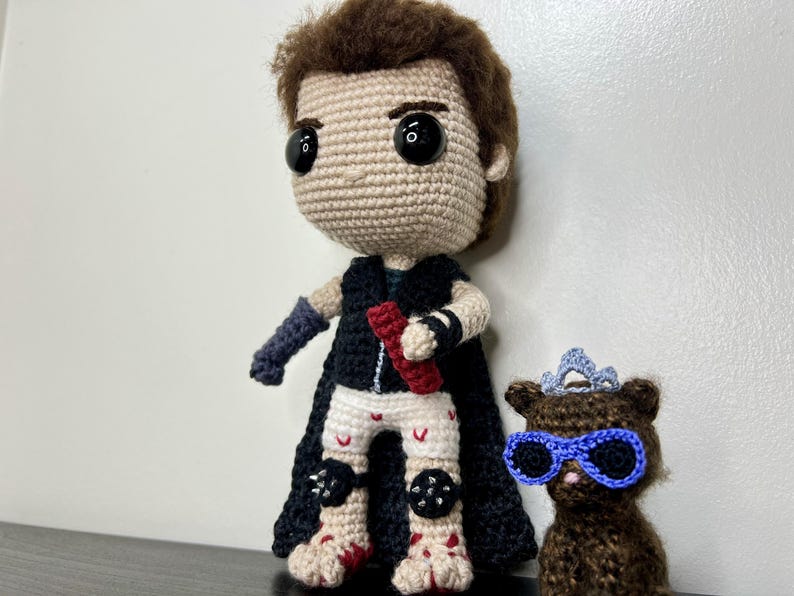 May include: Crocheted doll of a character with black cape, black and white pants, and a red object. Next to it is a crocheted bear wearing blue glasses and a crown. The dolls are handmade and have large black eyes.