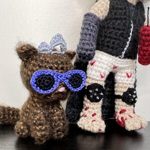 May include: Crocheted figures of a brown bear wearing blue sunglasses and a silver bow, and a figure in a black vest, white shorts with red accents, and black knee pads. The figure holds a red dynamite stick.