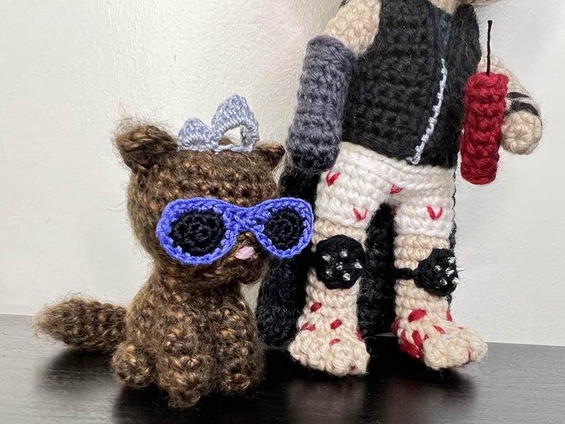 May include: Crocheted figures of a brown bear wearing blue sunglasses and a silver bow, and a figure in a black vest, white shorts with red accents, and black knee pads. The figure holds a red dynamite stick.