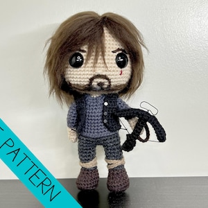 May include: A handmade crochet doll with brown hair, a beard, and a small wound. The doll wears a blue outfit and holds a black crossbow. The words "PDF PATTERN" are printed on a teal diagonal stripe.