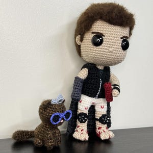 May include: Crocheted doll of a character with a black vest, gray arm sleeves, and white shorts with red accents. A small crocheted brown cat with blue glasses and a bow is next to the doll. The doll has brown hair and large black eyes.