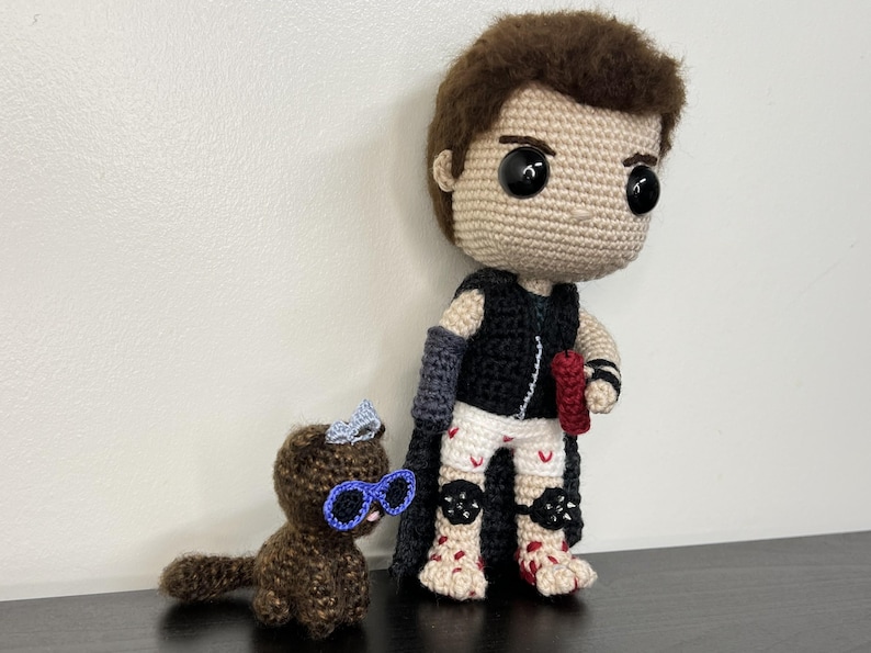 May include: Crocheted doll of a character with a black vest, gray arm sleeves, and white shorts with red accents. A small crocheted brown cat with blue glasses and a bow is next to the doll. The doll has brown hair and large black eyes.