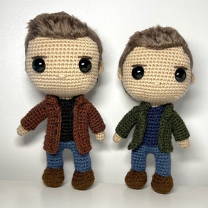 May include: Two crocheted figures of people wearing jackets and jeans. The figure on the left is wearing a brown jacket and the figure on the right is wearing a green jacket.