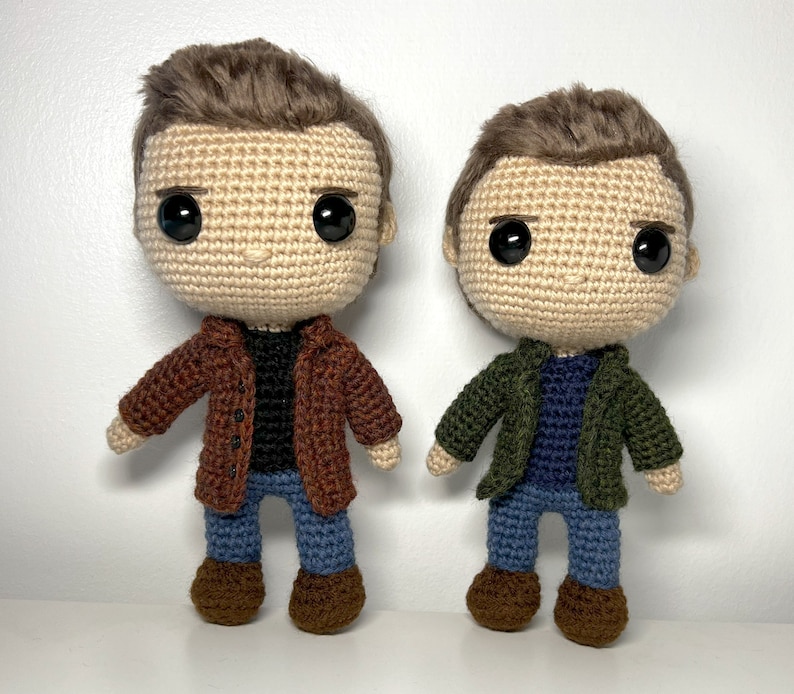 May include: Two crocheted figures of people wearing jackets and jeans. The figure on the left is wearing a brown jacket and the figure on the right is wearing a green jacket.