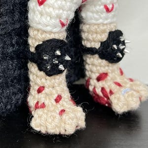 May include: Crocheted doll legs with black accents and silver spikes. The legs are cream-colored with red accents, and the doll has black crochet accents around the ankles. The doll's feet are also cream-colored with red accents.