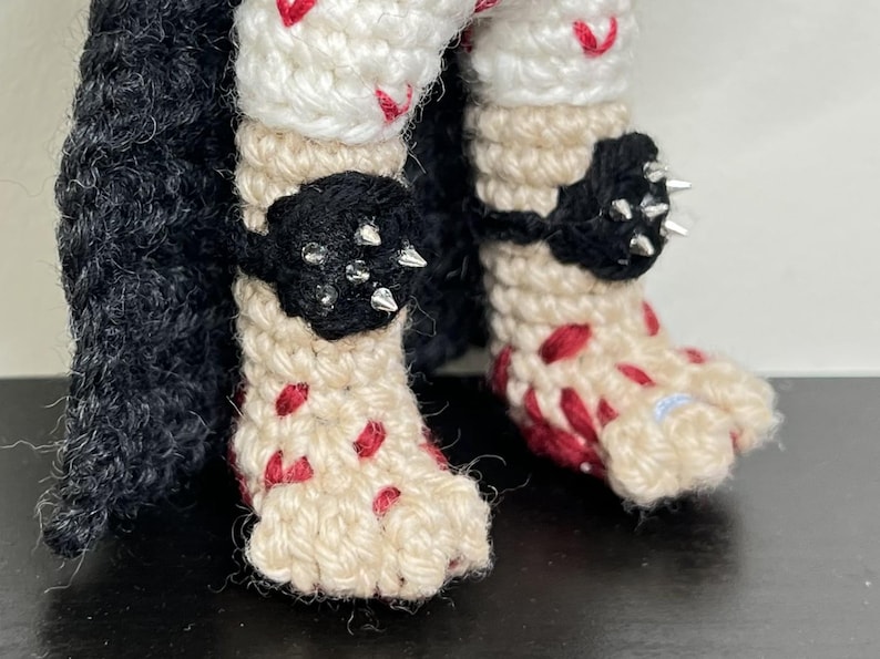 May include: Crocheted doll legs with black accents and silver spikes. The legs are cream-colored with red accents, and the doll has black crochet accents around the ankles. The doll's feet are also cream-colored with red accents.