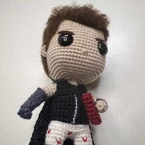May include: A crocheted doll with brown hair, large black eyes, and a tan face. The doll wears a black outfit with a cape, blue and gray arm sleeves, and white shorts with red accents. A red object is held in its right hand.