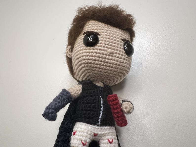 May include: A crocheted doll with brown hair, large black eyes, and a tan face. The doll wears a black outfit with a cape, blue and gray arm sleeves, and white shorts with red accents. A red object is held in its right hand.