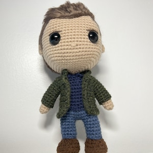 May include: Crocheted amigurumi doll with brown hair, a green jacket, a blue shirt, blue jeans, and brown shoes.