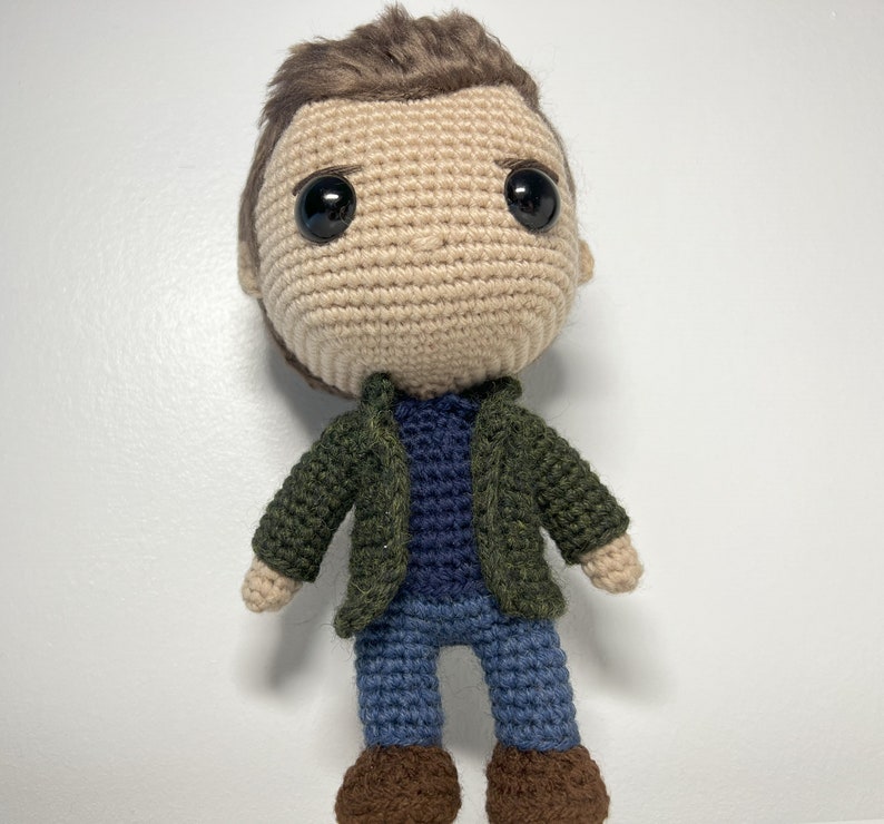 May include: Crocheted amigurumi doll with brown hair, a green jacket, a blue shirt, blue jeans, and brown shoes.