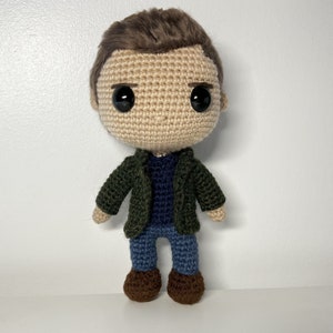 May include: Crocheted amigurumi doll with brown hair, a green jacket, a blue sweater, blue jeans, and brown shoes.