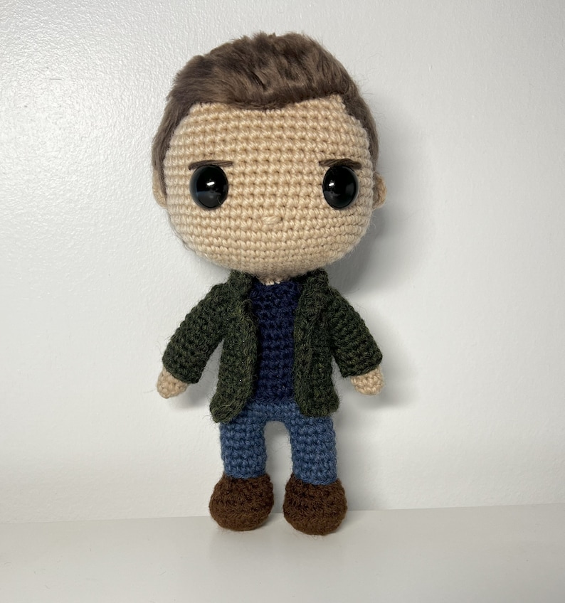 May include: Crocheted amigurumi doll with brown hair, a green jacket, a blue sweater, blue jeans, and brown shoes.