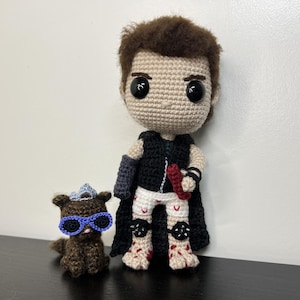 May include: Crocheted doll of a character with brown hair, black cape, and white shorts. The doll is holding a red object. A small crocheted animal with blue sunglasses sits beside the doll. The dolls are handmade.