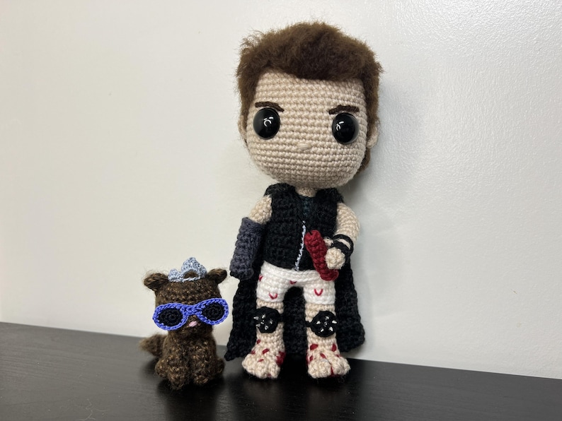 May include: Crocheted doll of a character with brown hair, black cape, and white shorts. The doll is holding a red object. A small crocheted animal with blue sunglasses sits beside the doll. The dolls are handmade.