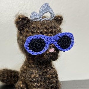 May include: Crocheted brown bear wearing blue sunglasses and a light blue crown. The bear has black eyes and a pink nose. The handmade bear is a unique, whimsical item.
