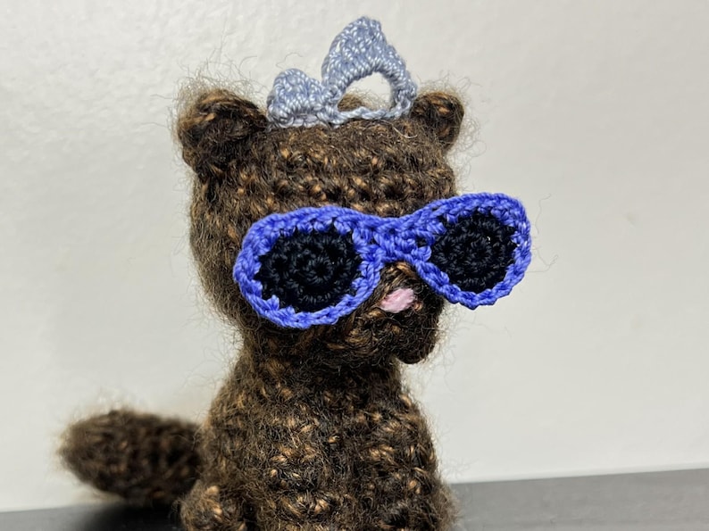 May include: Crocheted brown bear wearing blue sunglasses and a light blue crown. The bear has black eyes and a pink nose. The handmade bear is a unique, whimsical item.