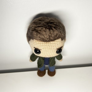 May include: Crocheted plush toy of a person with brown hair, wearing a green jacket, blue jeans, and brown shoes.