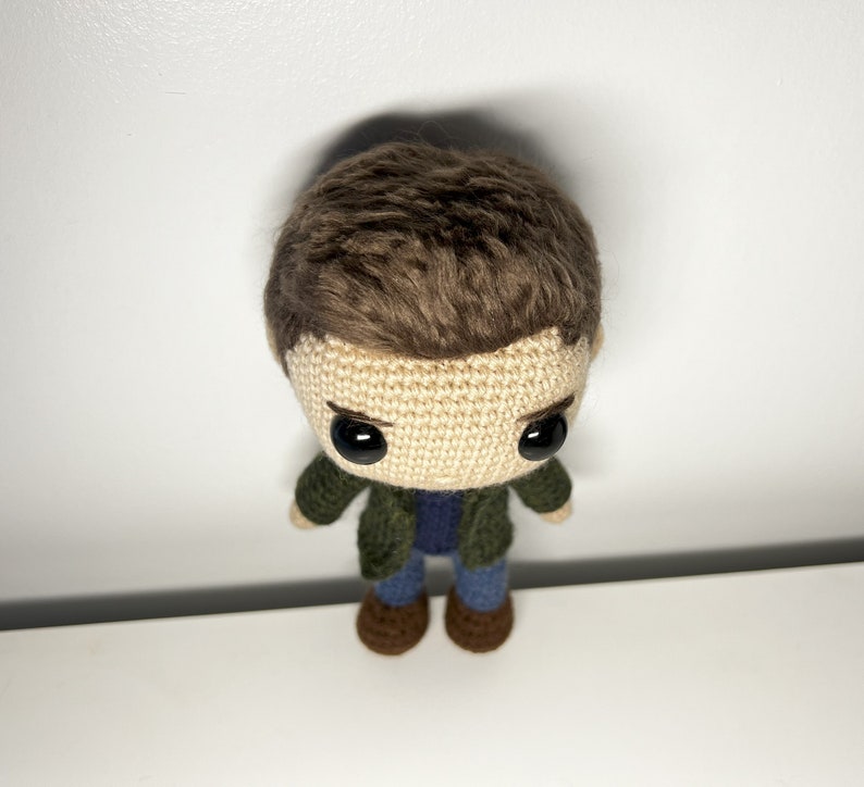 May include: Crocheted plush toy of a person with brown hair, wearing a green jacket, blue jeans, and brown shoes.