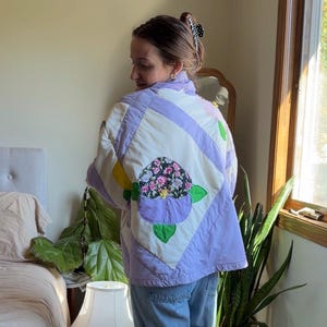 May include: A quilted jacket featuring a patchwork design in shades of lavender and white. The back displays a large floral appliqué with a black background, pink and white flowers, and green leaves, accented by lavender and green fabric petals.