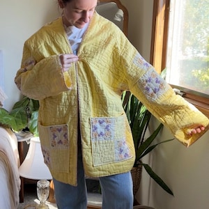 May include: A long-sleeved, open-front jacket crafted from yellow quilted fabric. The garment features two large patch pockets on the front, each accented with square floral patches in shades of purple and orange. The edges are finished with trim.