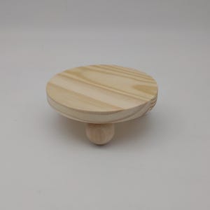 May include: A small, circular wooden riser featuring a light-colored, natural wood grain finish. The platform is supported by a single, centered spherical wooden foot, creating a low-profile stand with a smooth, unfinished surface texture.