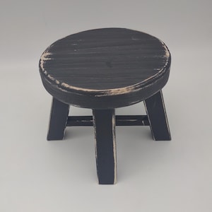 May include: A small, round wooden stool featuring a distressed black finish with visible light-colored wood grain showing through the edges. The stool is supported by three sturdy, angled legs connected by horizontal crossbars for stability.
