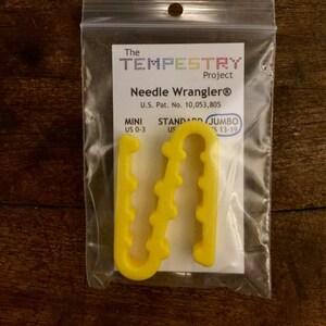 May include: A yellow plastic needle wrangler tool for knitting and crocheting. The tool is designed to hold needles in place while working on a project. The packaging includes the text "The TEMPESTRY Project Needle Wrangler® U.S. Pat. No. 10,053,805 MINI STANDARD JUMBO US 0-3 US 'S 13-19".
