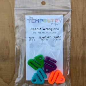 May include: A set of six colorful plastic needle threaders in a clear plastic bag. The threaders are labeled "Mini US 0-3", "Standard US 4-11", and "Jumbo US 13-19". The bag also includes the text "The wyvwywww Project Needle Wrangler® U.S. Pat. No. 10,053,805".