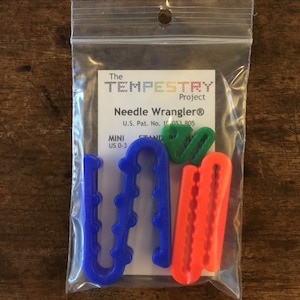 以下が含まれることがあります： タペストリー針のクラフト用のカラフルなプラスチック製の針ホルダーのセットが入った透明なビニール袋。針ホルダーは青、緑、オレンジ、赤です。袋には、「The Tapestry Project Needle Wrangler® U.S. Pat. No. 10,053,805 MINI US 0-3 STAND」と書かれたラベルが付いています。