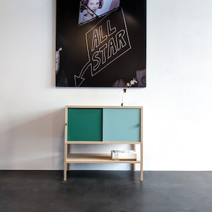 Puede incluir: Un armario de madera clara con dos puertas correderas. Las puertas están pintadas en dos tonos de verde. El armario tiene un estante y está situado en un suelo gris oscuro. Una silla negra es visible en el fondo.