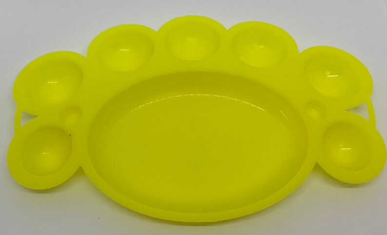 May include: A yellow plastic paint palette with seven individual wells for mixing paint. The palette is shaped like a paw print.