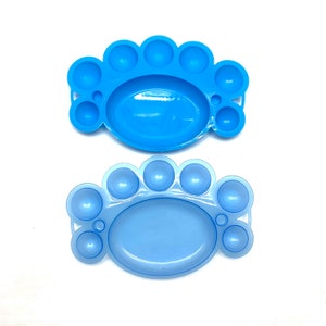 May include: Two blue plastic paint palettes with multiple circular wells. The palettes are designed for holding and mixing paint.