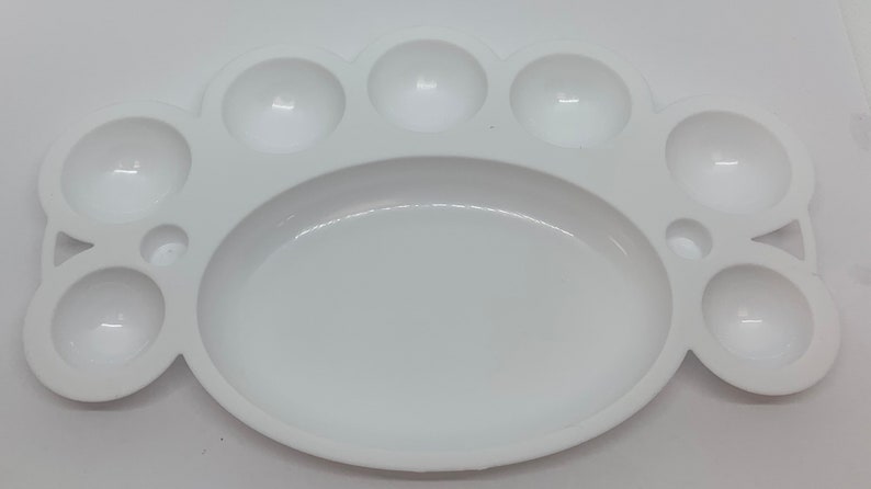 May include: White plastic paint palette with nine circular wells for mixing paint. The palette has a large oval area in the center.