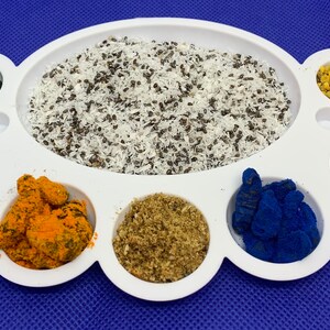 May include: A white ceramic palette with eight compartments, each filled with a different colored powder or ingredient. The compartments contain green, gray, orange, brown, blue, yellow, and white powders.