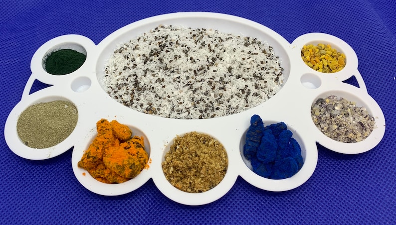 May include: A white ceramic palette with eight compartments, each filled with a different colored powder or ingredient. The compartments contain green, gray, orange, brown, blue, yellow, and white powders.